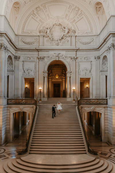 A couple holds hands while walking up grand marble stairs in an ornate, elegant building, reminiscent of wedding portraits with a Colorado mountain backdrop.