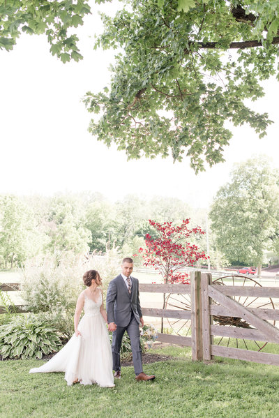 Bride and Groom holding hands and walking along wooden fence at farm on their sunny wedding day.