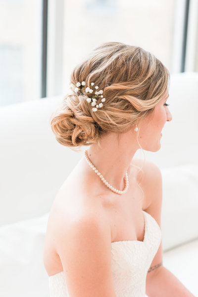 A bride sitting on a white couch wearing a strapless dress looking down with the focus on her updo hair with babies breath at the Glass Box at 230