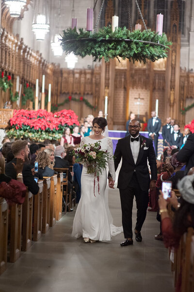 Bride in long sleeve wedding dress with lace detail holding. dark winter bouquet holding hands with a grooms wearing black tux leaving duke chapel durham nc