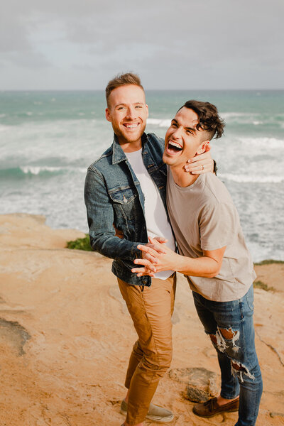 A blonde man and his brunette husband laugh together on a beach