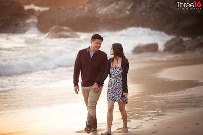Engaged couple walks along Thousand Steps Beach in Laguna Beach holding hands and smiling at each other