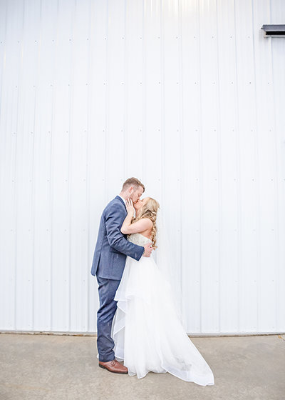 Bride and groom kiss by white barn on wedding day