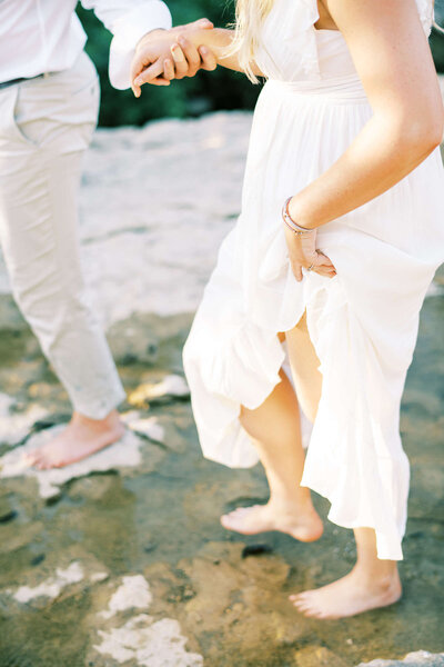 Engaged couple dressed in white holds hands as they cross a river
