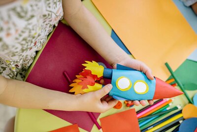 A child is holding a colorful, handmade paper rocket craft with red, yellow, and blue elements, surrounded by various art supplies and colored paper on a table.