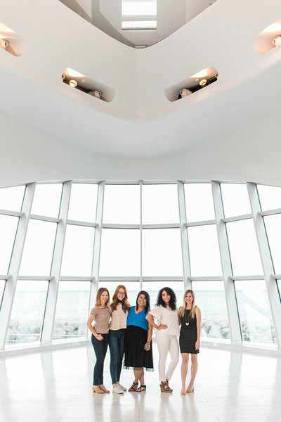 five women standing together smiling in a bight modern art museum building with a lake in the background