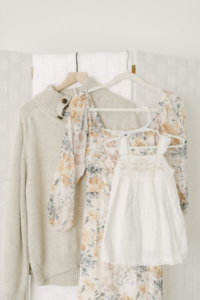 A picture of a family wardrobe selection, a beige sweater for dad, a peach and navy floral dress for mom and a white tank dress with pink embroidery for baby