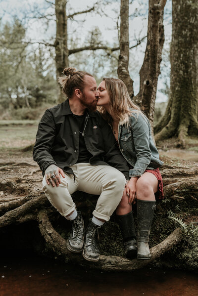 Beautiful couple kiss whilst sitting on large tree vines by the river in New Forest