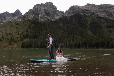 A bride sits on a paddleboard in the middle of a lake while her new husband paddles her around.