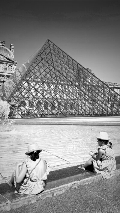 Two little girls sitting with feet in fountain at the louvre france with glass pyramid in the background.
