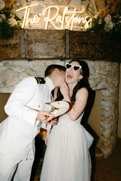 Bride and groom eating cake in front of their last name neon sign. Bride in heart shaped sunglasses while groom is kissing her neck