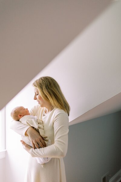 Mom holding newborn baby with window light streaming into the nursery
