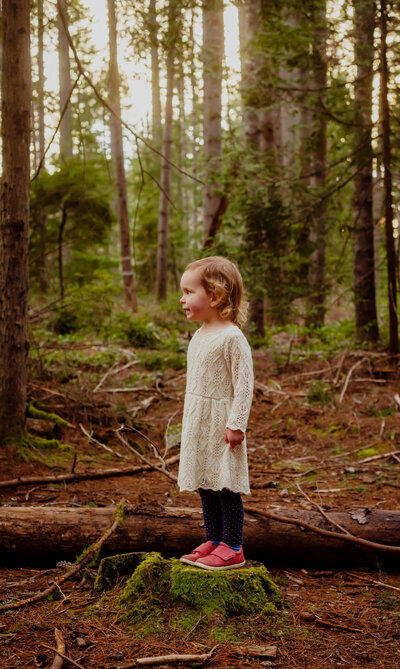 Toddler girl in a white knit dress and pink shoes standing on a mossy tree stump in the woods looking off into the distance