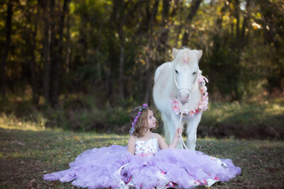 girl-in-purple-floral-couture-dress-holding-unicorn-rein-with-unicorn-wearing-floral-necklace-in-dalworthington-gardens