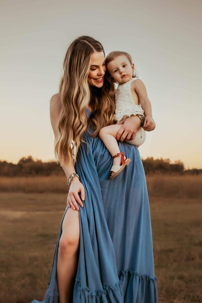 A Macon family photographer captures a woman in a blue dress holding her baby in a field at sunset.