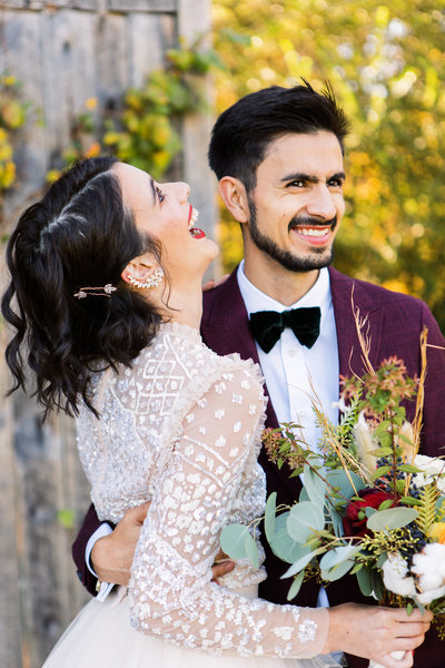 Young wedding couple laughing together with bride wearing diamon ear cuff and groom in a maroon suit.