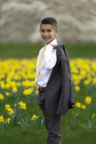 A young boy walks through a field of daffodils carrying his grey suit jacket over his shoulder as directed by a New Jersey Communion Portrait Photographer