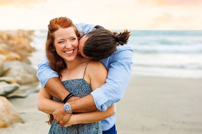Man holding his wife in a bear hug posing in front of the rock jetty at Oceanside Harbor Beach