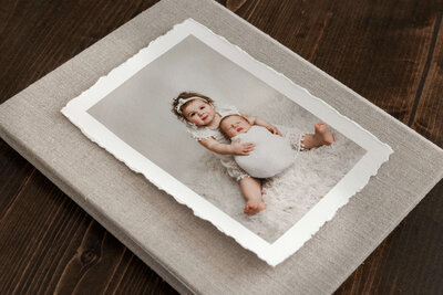 print of older sister holding her newborn brother who is swaddled laying on top of a tan folio