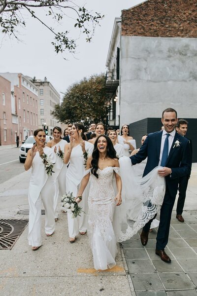 Groom carries brides dress as they walk to the ceremony, bridesmaids in white jumpsuits
