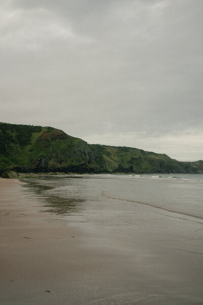 A quiet shoreline under an overcast sky, with soft waves meeting the sand and green cliffs stretching along the coast.