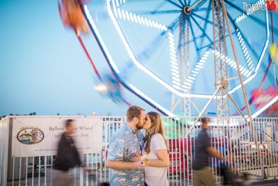 Engaged couple share a kiss in front of the Ferris Wheel at the Balboa Fun Zone in Newport Beach