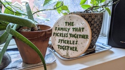 An embroidery hoop saying "The family that pickles together stickles together," surrounded by plants. Embroidery and photo by Hannah de Keijzer.