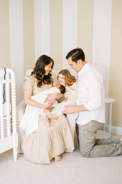 Mom holds newborn baby boy while big sister gives a kiss in neutral nursery with jenny lind crib during newborn session in their home