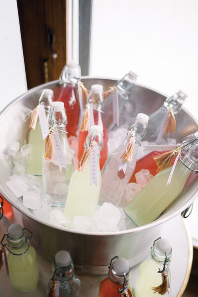 Pink and yellow themed baby shower drink bottles in a silver ice bucket, each labeled with drink names and adorned with matching tassels, creating a festive and stylish refreshment display.