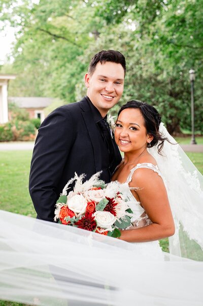 Couple smiling at the camera outside of Oaklands Mansion on their wedding day, her flowers are cream and red and her veil is blowing in the wind
