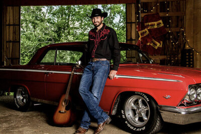 Country music portrait Chance Moore standing next to red classic car guitar leaning on car door beside him parked inside barn trees through entrance behind him