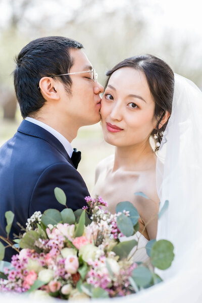 Chinese couple's micro wedding in Lubbock TX.  Bride is looking at the camera while groom is kissing her cheek with his eyes closed.