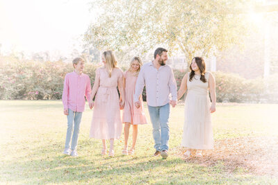 A family of five dressed in hues of neutrals and pink hold hands and laugh while walking through a field.