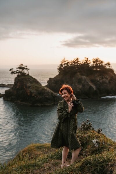 Woman in a green dress stands barefoot on a cliff by the ocean at sunset, with rocky islands behind her—an inspiring scene from an outdoor shoot during photo retreat weekend.