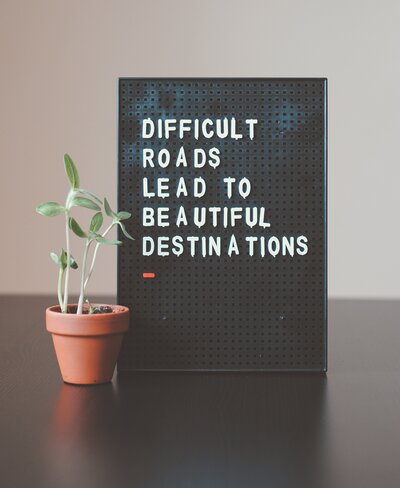 Image of a framed message that reads "Difficult Roads Lead to Beautiful Destinations" next to a small potted plant