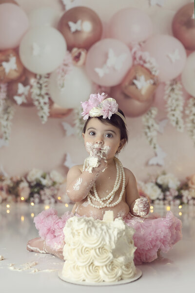A toddler girl in a pink tutu and floral headband sits covered in cake for her first birthday cake smash in a studio