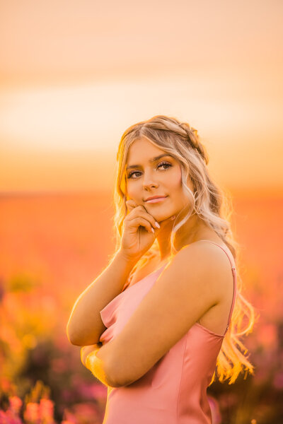 blonde senior girl with long wavy hair standing in a field of bright pink flowers at sunset