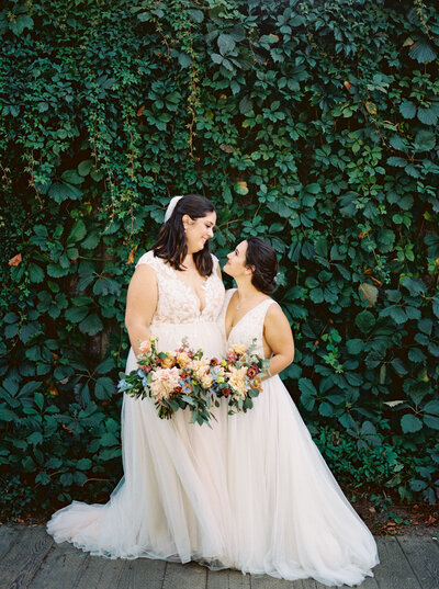 Two brides wearing white flowing gowns holding bouquets looking at each other