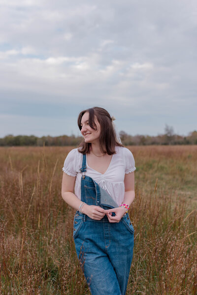 A League City Texas senior poses in a brown field in overalls with a white top and pink friendship bracelets