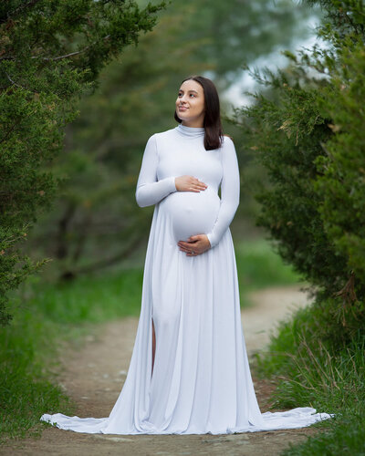 A pregnant woman stands in the forest on a path, wearing a long white maternity gown, looking off in the distance.