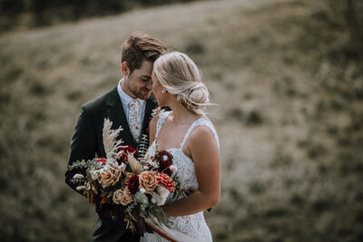 Bride and groom embracing, holding gorgeous burgundy and taupe fall bouquet by Fiore Fine Events, wedding planner in Calgary, Alberta, featured on the Brontë Bride Vendor Guide.