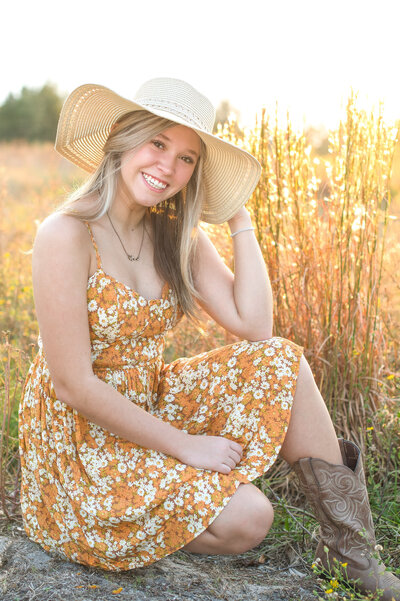 High school senior girl in sun hat and cowboy boots sitting in a field.