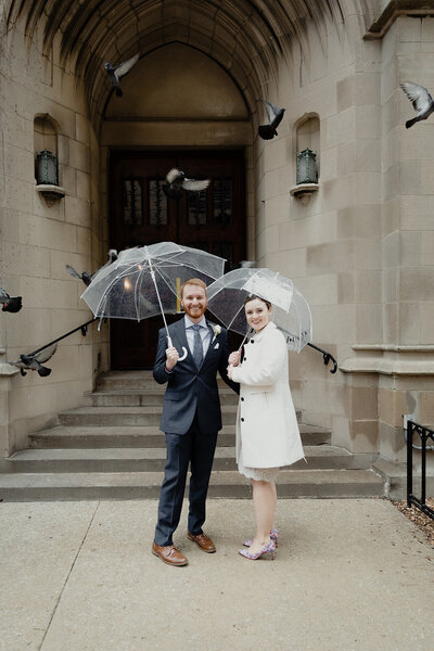couple stands outside of church after their ceremony with clear umbrellas and pigeons flying overhead