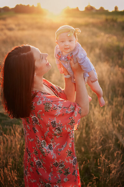 mom in red shirt holding baby smiling in field at sunset in minneapolis