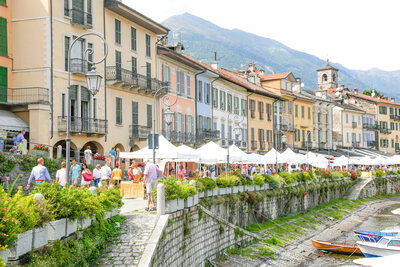 A vibrant street market in a European town features colorful buildings with balconies lining a canal. People shop at stalls under white tents, and small boats are docked by the water. Mountains are visible in the background.