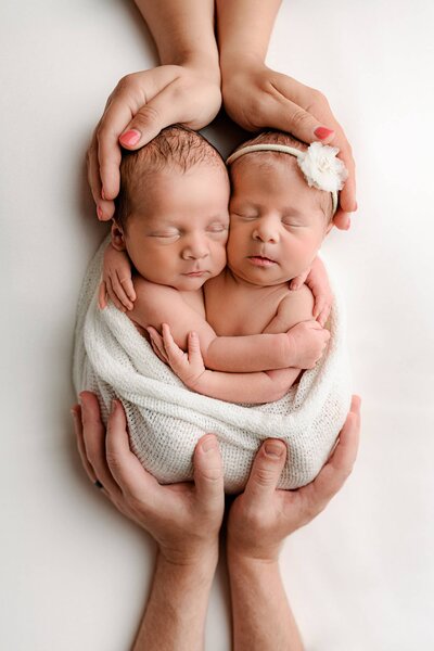 boy girl twin newborns wrapped in white together with parents hands at top and bottom 