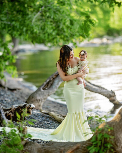 A young mother is standing with her baby daughter, wearing a pale yellow gown by the lake