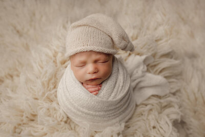 A newborn boy wrapped in all white sleeping in a sitting position