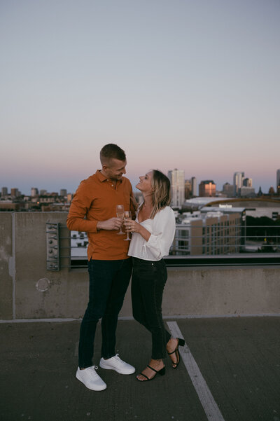 Engagement photos taken on a rooftop in Milwaukee, WI at sunset with the man and woman toasting and cheersing their champagne, looking at eachother smiling