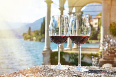 Two glasses of red wine on a stone ledge, overlooking a scenic lake with mountains and villas in the background. Sunlight creates a warm glow, highlighting the elegant archways and lush greenery surrounding the tranquil setting.